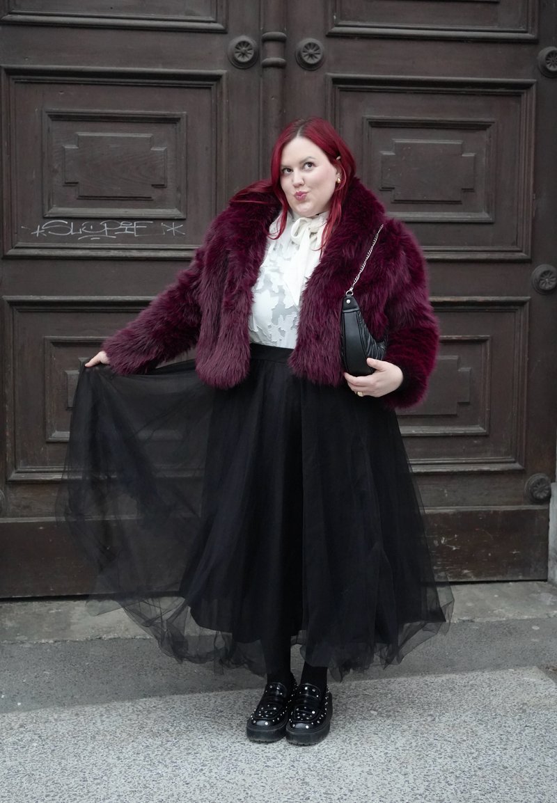 Ženska rdečelasa, oblečena v bordo bundico iz umetnega krzna, belo bluzo in črno tilasto krilo, drži krilo in črno torbico.