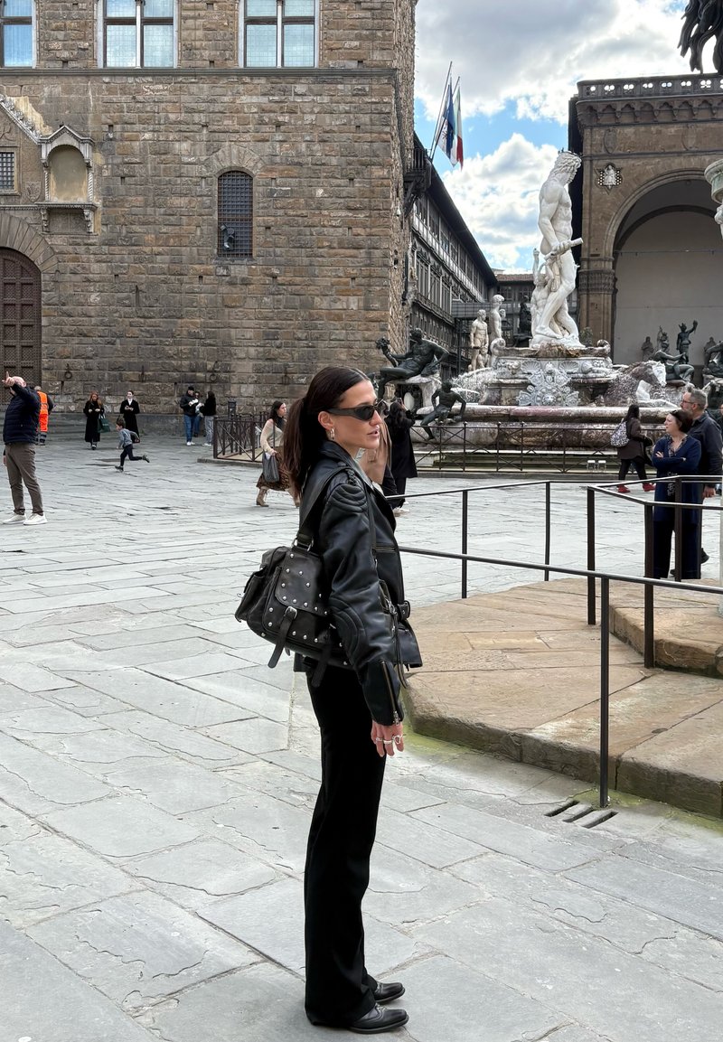 Žena v čiernej koženej bunda a slnečných okuliaroch stojaca pri renesančných sochách a historickej kamennej budove na vonkajšom námestí s ďalšími návštevníkmi.