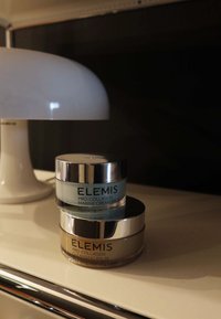 Dwa ustawione jeden na drugim słoiki kosmetyków Elemis Pro-Collagen na blacie obok białej, zaokrąglonej lampy stołowej w słabym świetle.