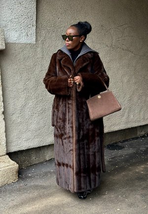 Mujer que lleva un abrigo largo de piel marrón oscuro, gafas de sol, sosteniendo un bolso marrón, de pie frente a una pared gris texturizada.