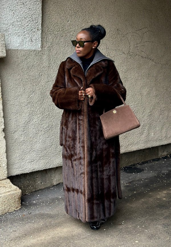 Sieviete, tērpusies garā tumši brūnā kažokā, saulesbrillēs, turot brūnu rokassomiņu, stāv pret teksturētu pelēku sienu.