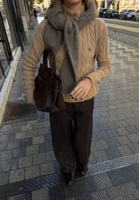 Beige kabelstrikket genser med høy krage, grått skjerf drapert rundt halsen, brun håndveske, vide mørke bukser og brune sko på brosteinsbelagt gate.