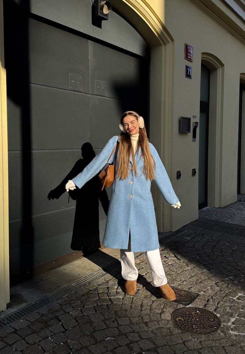 Woman in blue coat, white pants, and earmuffs stands smiling with arms outstretched against dark garage door in sunlight.