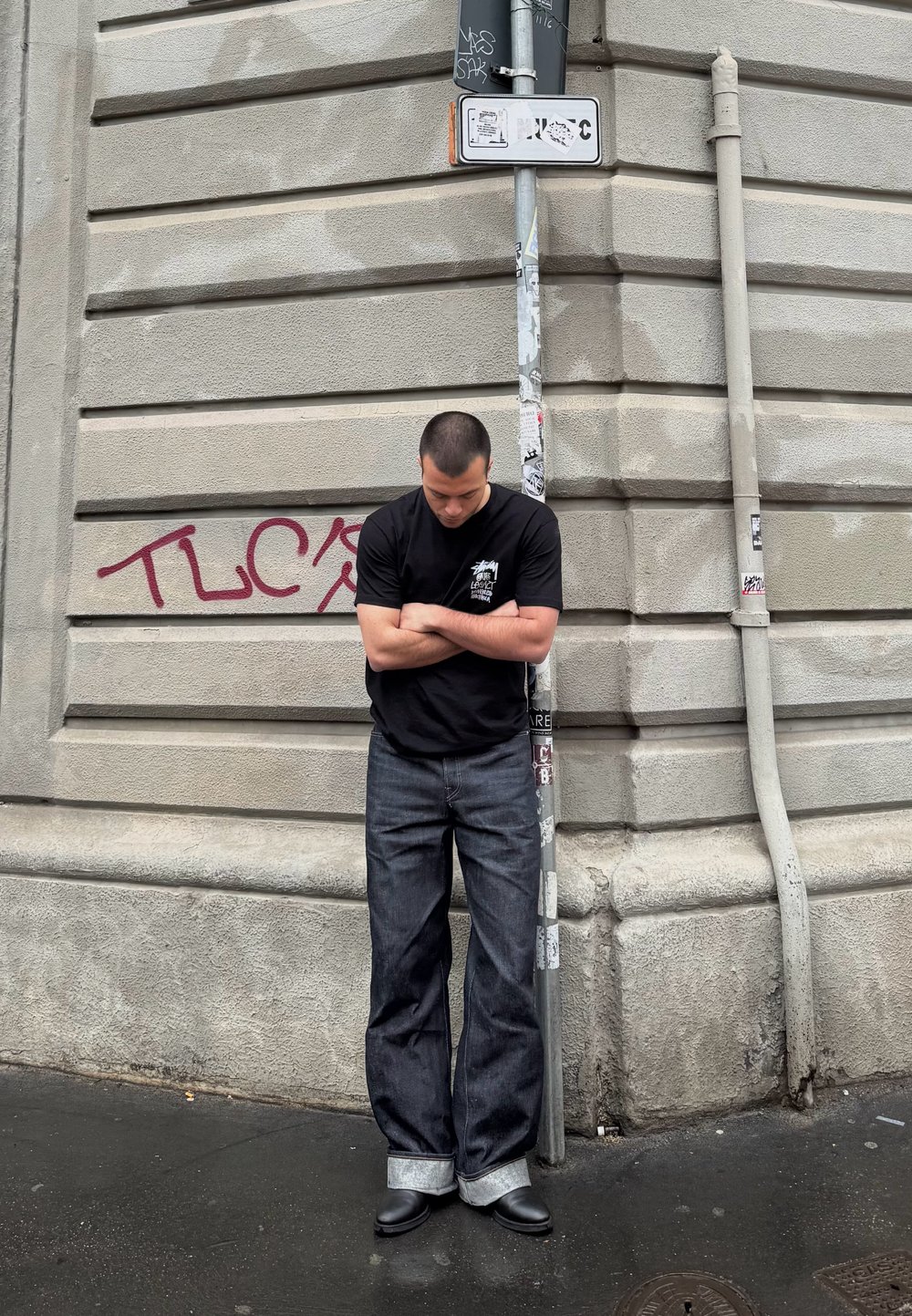 Črna majica, široke temno modre kavbojke s manšetami in črne gležnjarje, stoji ob teksturirani bež stavbi z grafiti.