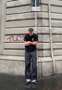 Črna majica, široke temno modre kavbojke s manšetami in črne gležnjarje, stoji ob teksturirani bež stavbi z grafiti.