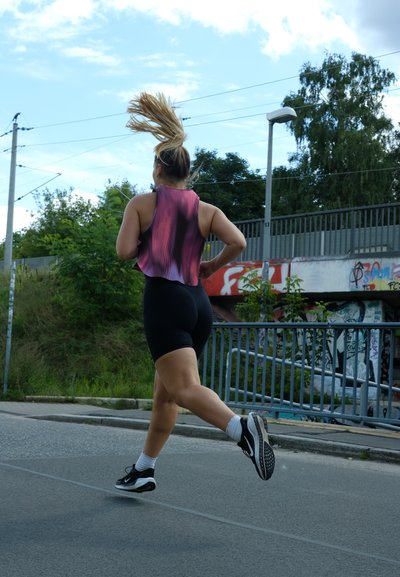 Corredor con una camiseta sin mangas rosa y negra y pantalones cortos negros, vistiendo zapatillas de correr negras con suelas blancas. Entorno al aire libre soleado con vegetación.