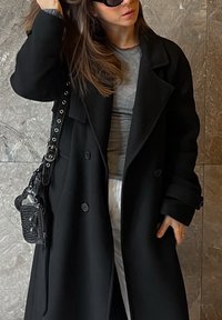 Lange zwarte jas met grote revers, dubbele knopen, gedragen over een grijs hemd. Zwarte handtas met een gestructureerde afwerking. Strak, modern ontwerp.