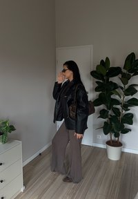 Žena v černé kožené bundě a slunečných brýlích pózuje v místnosti s dřevěnou podlahou, hnědými kalhotami a dvěma zelenými květináči poblíž.