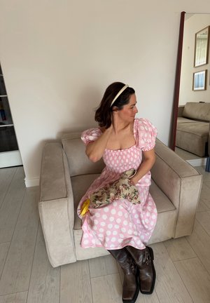 Mujer con vestido rosa de lunares y botas marrones sentada en un sillón beige, sosteniendo una bolsa de tela floral, mirando hacia su derecha en una sala de estar.