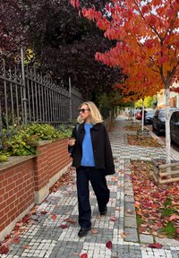 Must jakk sinise pulloveri, tumedate pükste ja päikeseprillidega. Inimene hoiab jalutades lehtedega kaetud munakivisillutisel jooki.