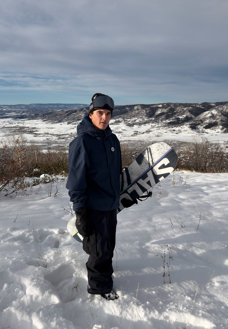 Planche de snowboard avec un design graphique noir et blanc, tenue par une personne portant une veste bleue, des gants noirs et des lunettes de protection, debout sur un terrain couvert de neige.
