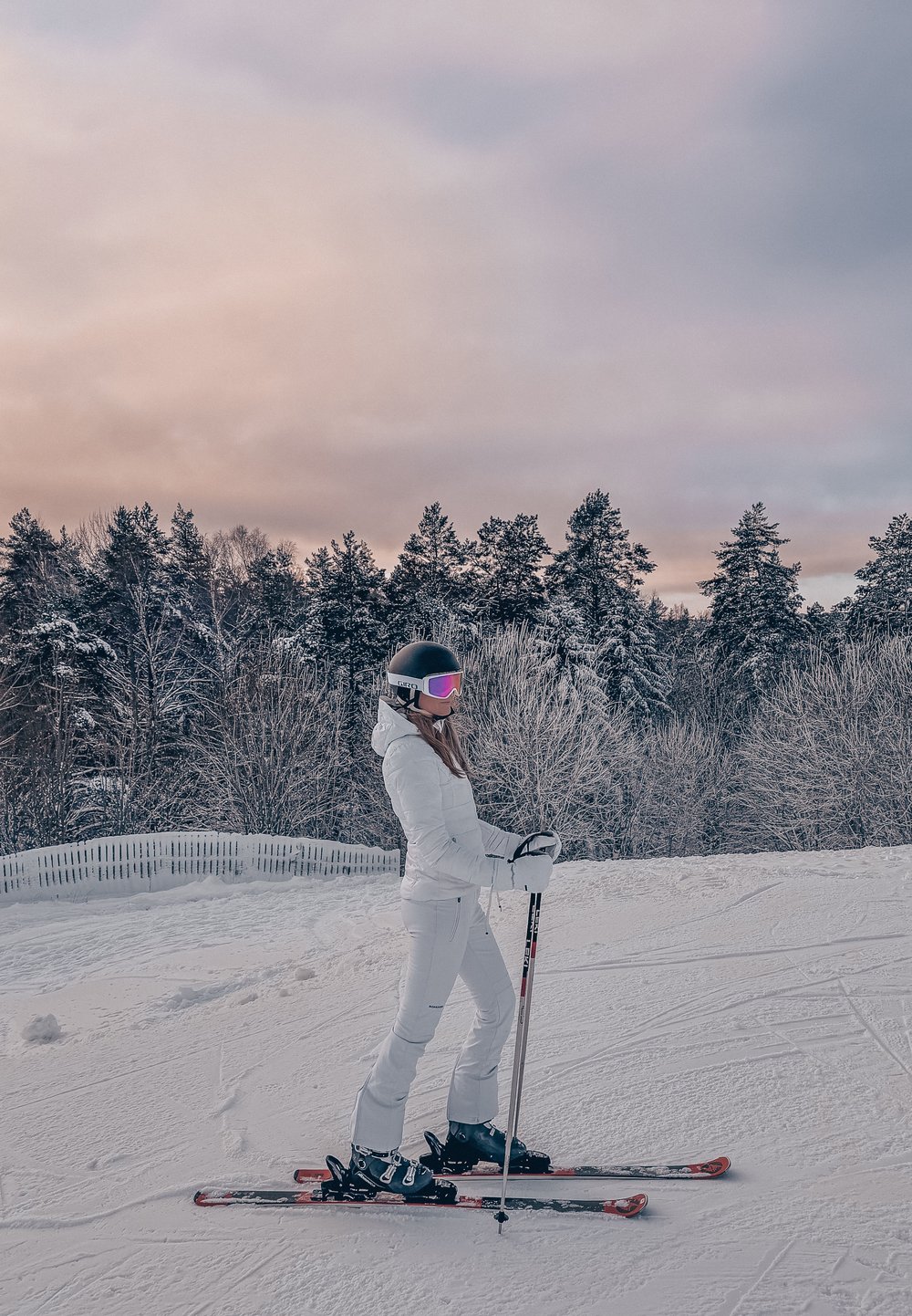 Personne en tenue de ski blanche et casque noir debout sur des skis dans un paysage enneigé avec des arbres couverts de neige et un ciel nuageux au coucher du soleil.