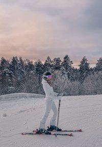 Personne en tenue de ski blanche et casque noir debout sur des skis dans un paysage enneigé avec des arbres couverts de neige et un ciel nuageux au coucher du soleil.