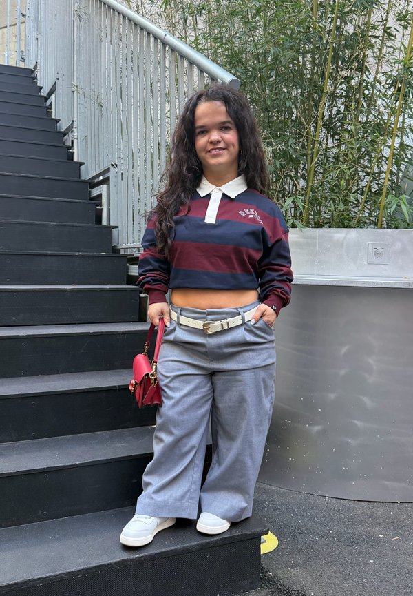 Una mujer está de pie en unos escalones exteriores negros, sostiene un bolso rojo y lleva una camiseta de rayas, unos pantalones grises holgados y unas zapatillas blancas.