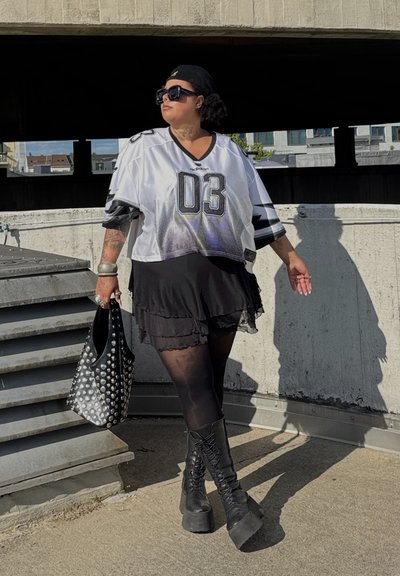 Camiseta de malla blanca con detalles negros y el número 03, superpuesta sobre una falda negra. Botas de plataforma negras con cordones y un bolso de mano con tachuelas.