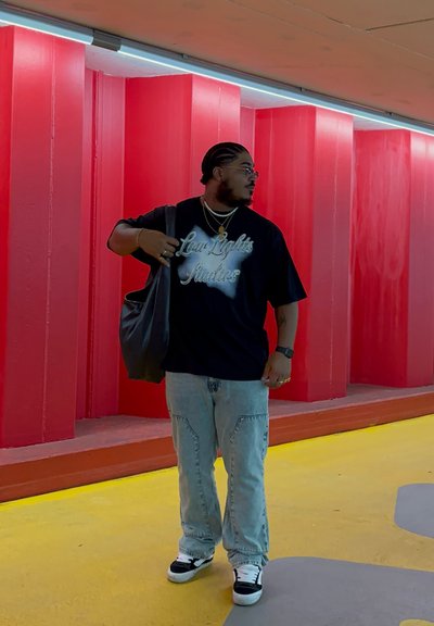 Camiseta negra con texto plateado "Derechos Legales", jeans azules claros y zapatillas negras y blancas. Sostiene una bolsa de mano de color oscuro. Fondo de pared roja.
