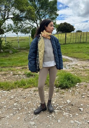 Chaqueta acolchada azul y verde sobre un suéter gris, camisa clara y leggings beige. Botines marrones, bufanda estampada y fondo natural al aire libre.