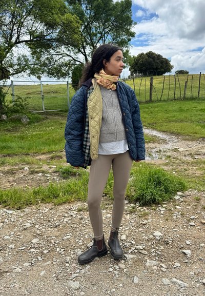 Chaqueta acolchada azul y verde sobre un suéter gris, camisa clara y leggings beige. Botines marrones, bufanda estampada y fondo natural al aire libre.