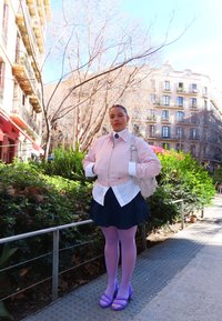 Jeune personne en veste rose, chemise blanche, jupe marine, collants lavande et sandales violettes, debout sur le trottoir d'une ville près de la végétation et des bâtiments.