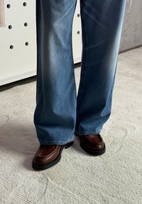 Persona con jeans a gamba larga blu e mocassini in pelle marrone scuro in piedi su un pavimento di moquette chiara all'interno.