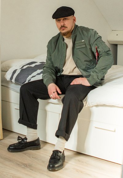 Chaqueta bomber verde oliva con acabado texturizado y múltiples bolsillos, combinada con pantalones negros y mocasines negros. Calcetines finos beige visibles.