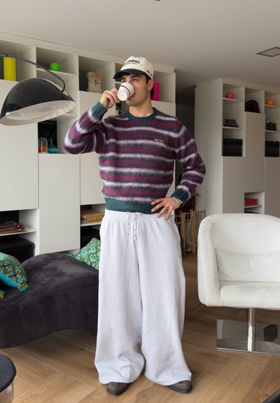Joven vestido con suéter a rayas y pantalones anchos blancos bebe de una taza pequeña en una sala moderna con estanterías blancas.