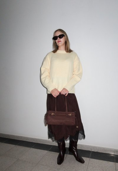 Suéter crema, falda de ante marrón con dobladillo de encaje, bolso marrón con detalles dorados, botas de cuero marrón hasta la rodilla y gafas de sol oscuras.