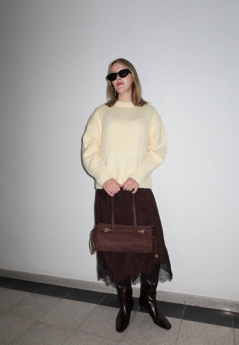 Pull crème, jupe en suède marron avec ourlet en dentelle, sac à main marron avec accents dorés, bottes en cuir marron montant jusqu'au genou, et lunettes de soleil foncées.