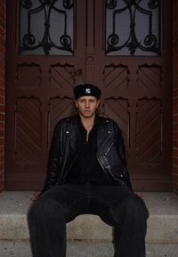 Een persoon die een zwarte leren jas en een baret draagt, zit op stenen trappen voor sierlijke dubbele houten deuren met decoratieve patronen.