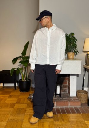 Hombre con camisa blanca abotonada, pantalones negros, zapatos marrones, gorra negra y gafas, de pie en una habitación con plantas y una chimenea.