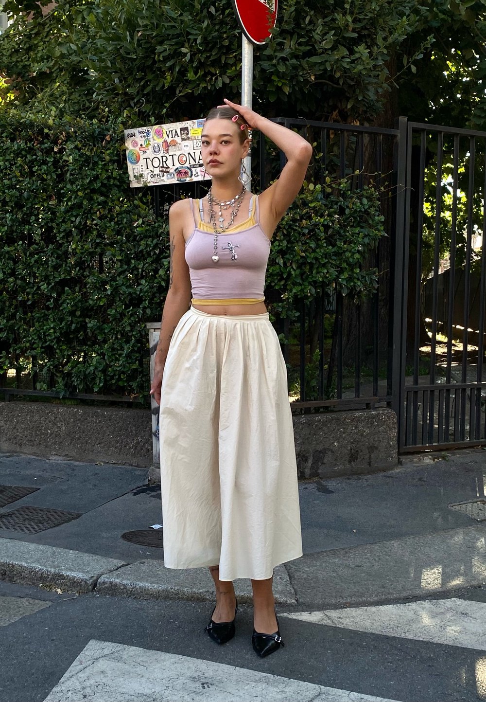 Top cropped rosa claro com detalhes em amarelo, combinado com uma saia midi plissada em creme. Ela usa sapatos pretos de bico fino e colares em camadas.