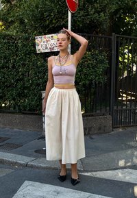 Top cropped rosa claro com detalhes em amarelo, combinado com uma saia midi plissada em creme. Ela usa sapatos pretos de bico fino e colares em camadas.