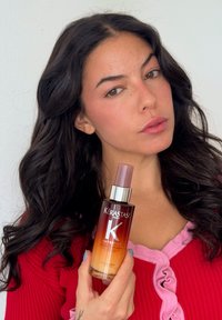 Kérastase Nutritive hårolie i en gennemsigtig flaske med en ombre-effekt, der har en sølvfarvet hætte og en lyserød spraydyse, holdt op mod en lys baggrund.