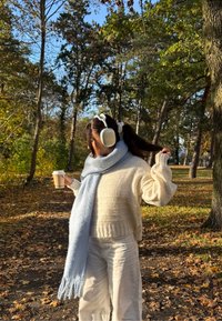 Beli pleteni pulover z svetlo modrim šalčkom, nosi velike bele slušalke in drži skodelico kave v gozdnatem območju z jesenskim listjem.