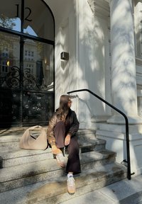 Καφέ δερμάτινο μπουφάν, μαύρο παντελόνι, μπεζ αθλητικά. Καθισμένος σε πέτρινες σκάλες δίπλα σε ένα άσπρο κτίριο με μαύρη γυάλινη πόρτα και διακοσμητικά στοιχεία.