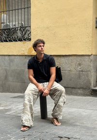 Egy fiatal férfi ül egy fekete fém oszlopon, fekete pólóinget és világos terepmintás nadrágot visel, papucsban, kezében fekete vászontáskával.