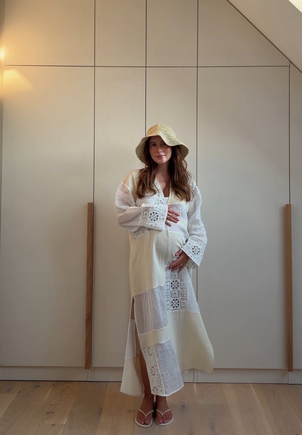 Femme portant une robe patchwork blanche et un chapeau de paille, debout pieds nus sur un sol en bois devant des placards beige, tenant son ventre.