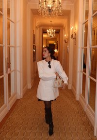 Cappotto bianco doppiopetto su un vestito nero, stivali neri alti fino al ginocchio e una borsa color crema. Ingresso interno con lampadari e tappeto decorato.
