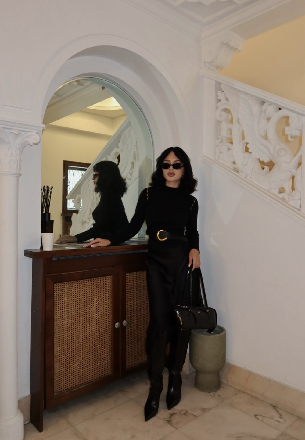 Mujer vestida completamente de negro con gafas de sol, de pie junto a un armario de madera y una escalera blanca ornamentada dentro de una habitación con suelo de mármol.