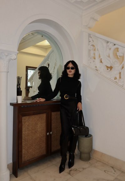 Mujer vestida completamente de negro con gafas de sol, de pie junto a un armario de madera y una escalera blanca ornamentada dentro de una habitación con suelo de mármol.