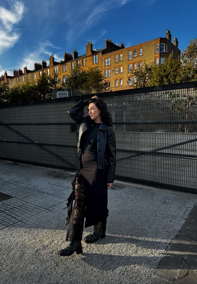 Chaqueta de cuero negra sobre un vestido negro ajustado con detalles de encaje. La modelo lleva botas con borde de encaje. Fondo urbano con arquitectura moderna.