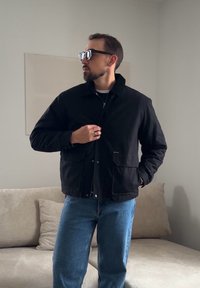 Homme portant une veste noire, un jean bleu et des lunettes de soleil, debout à l'intérieur près d'un canapé beige avec des coussins, regardant sur le côté.