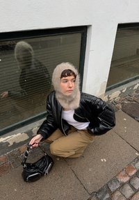 Personne portant un passe-montagne en tricot beige, une veste en cuir noire et un pantalon camel, accroupie sur le trottoir près d'un bâtiment, tenant un petit sac à main noir.