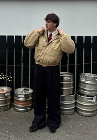 Bézs, túlméretezett dzseki magas gallérral, ezüst színű fémszerelvényekkel és zsebekkel. Fekete, széles nadrág és sötét lábbeli. Fém hordók a háttérben.