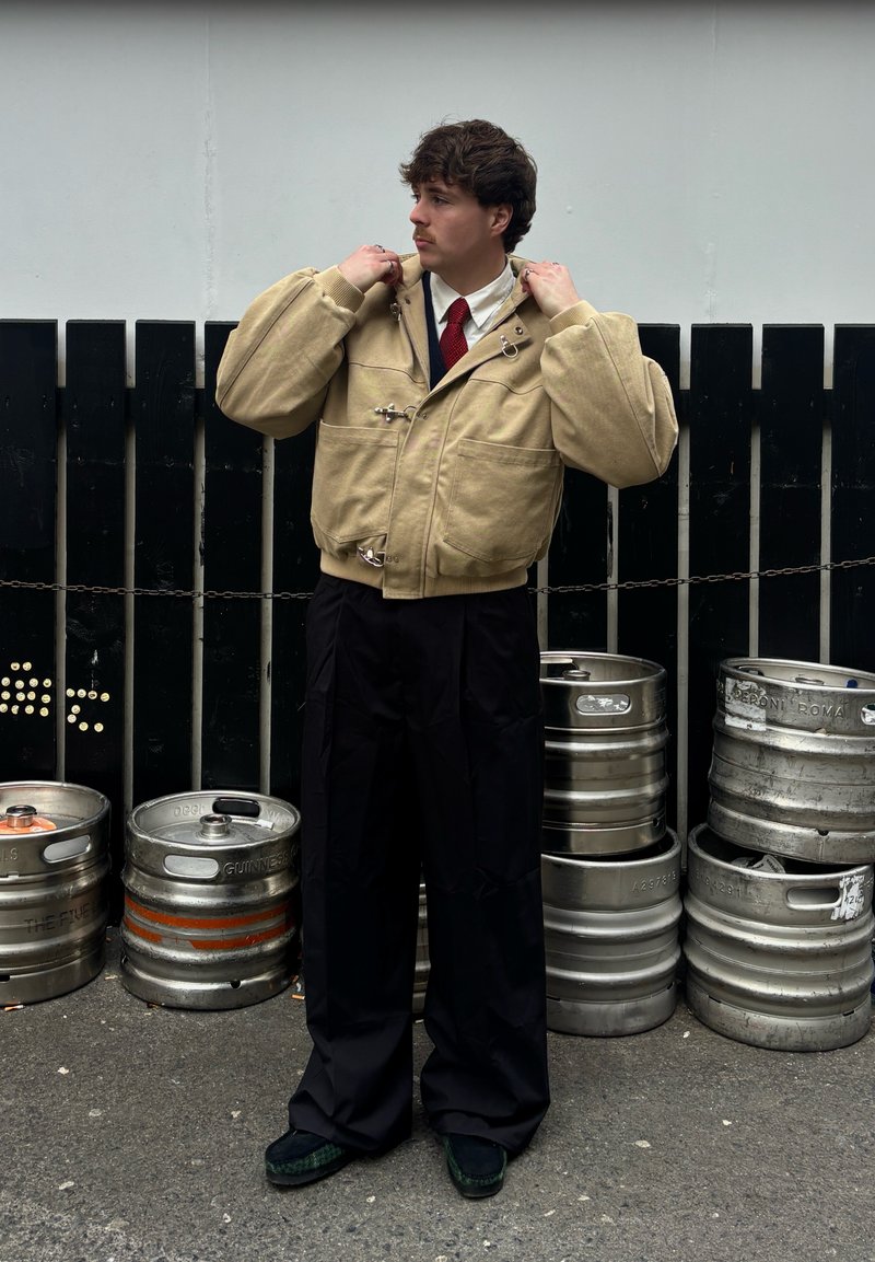 Veste beige surdimensionnée avec un col haut, dotée de ferrures argentées et de poches. Pantalon large noir et chaussures foncées. Fûts métalliques en arrière-plan.