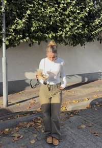 Hvit langermet topp, olivengrønne vide bukser, brune støvler. Holder en kald drink og solbriller, med falne blader på bakken.