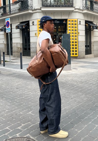 Bolso de viaje de cuero marrón con correas laterales, llevado sobre una camisa blanca y un chaleco beige, combinado con jeans de mezclilla oscura y zapatos beige.