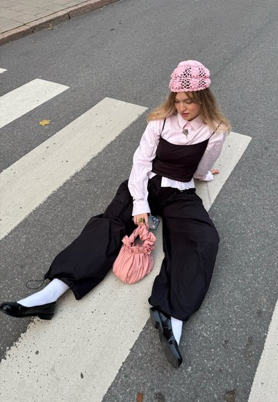 Mujer joven con gorro de punto rosa y blusa con un conjunto negro en capas se sienta en un paso de peatones sosteniendo una bolsa rosa y un teléfono.