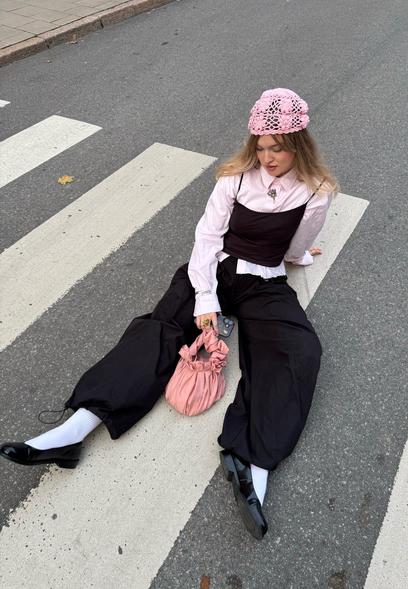 Young woman in pink knit hat and blouse with black layered outfit sits on a crosswalk holding a pink bag and phone.