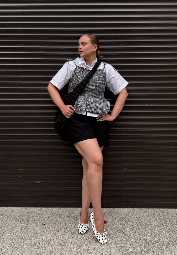 Jeune femme portant un chemisier blanc avec un haut à carreaux, un short noir, des chaussures à pois et un sac à épaule noir, debout devant un mur sombre.